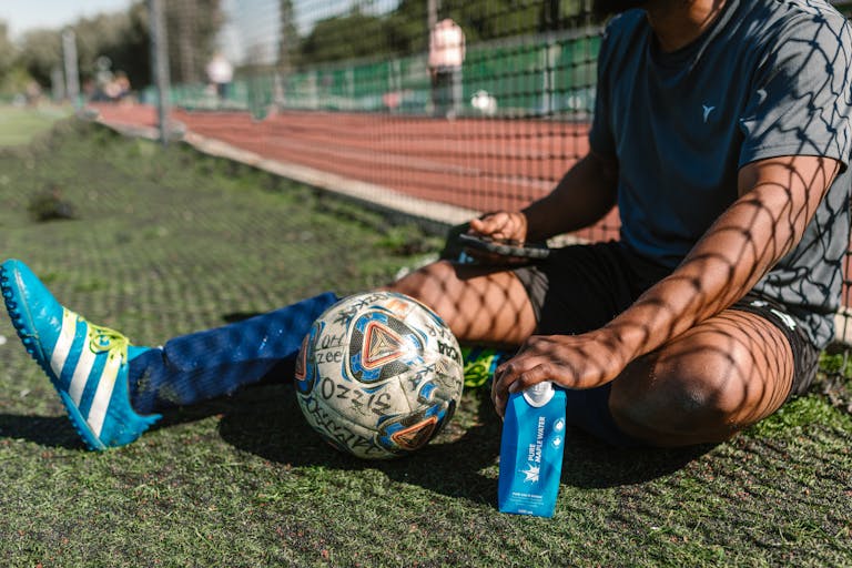 A soccer player takes a break, sitting on a grassy field with a soccer ball and water bottle.
