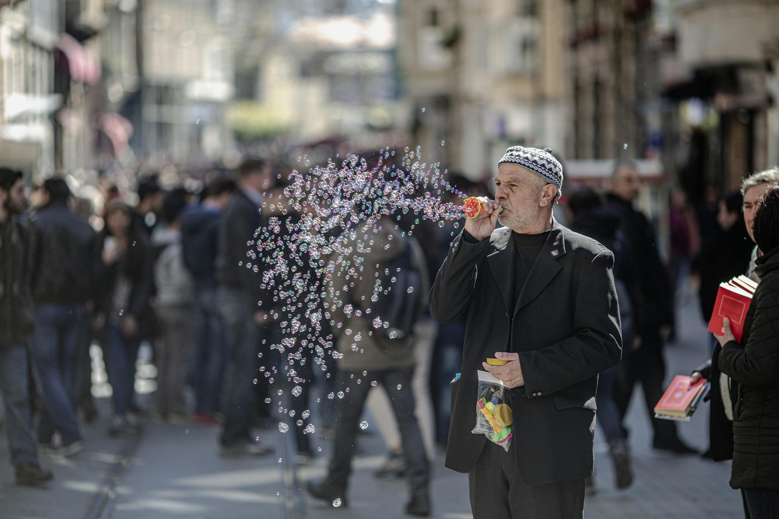 An elderly man blows bubbles on a crowded street in Istanbul, creating a whimsical and lively scene.
